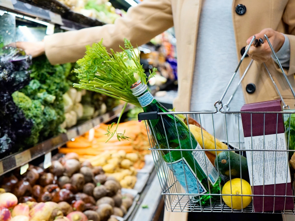 A person holding a basket full of groceries, grabbing something from the vegetables area.